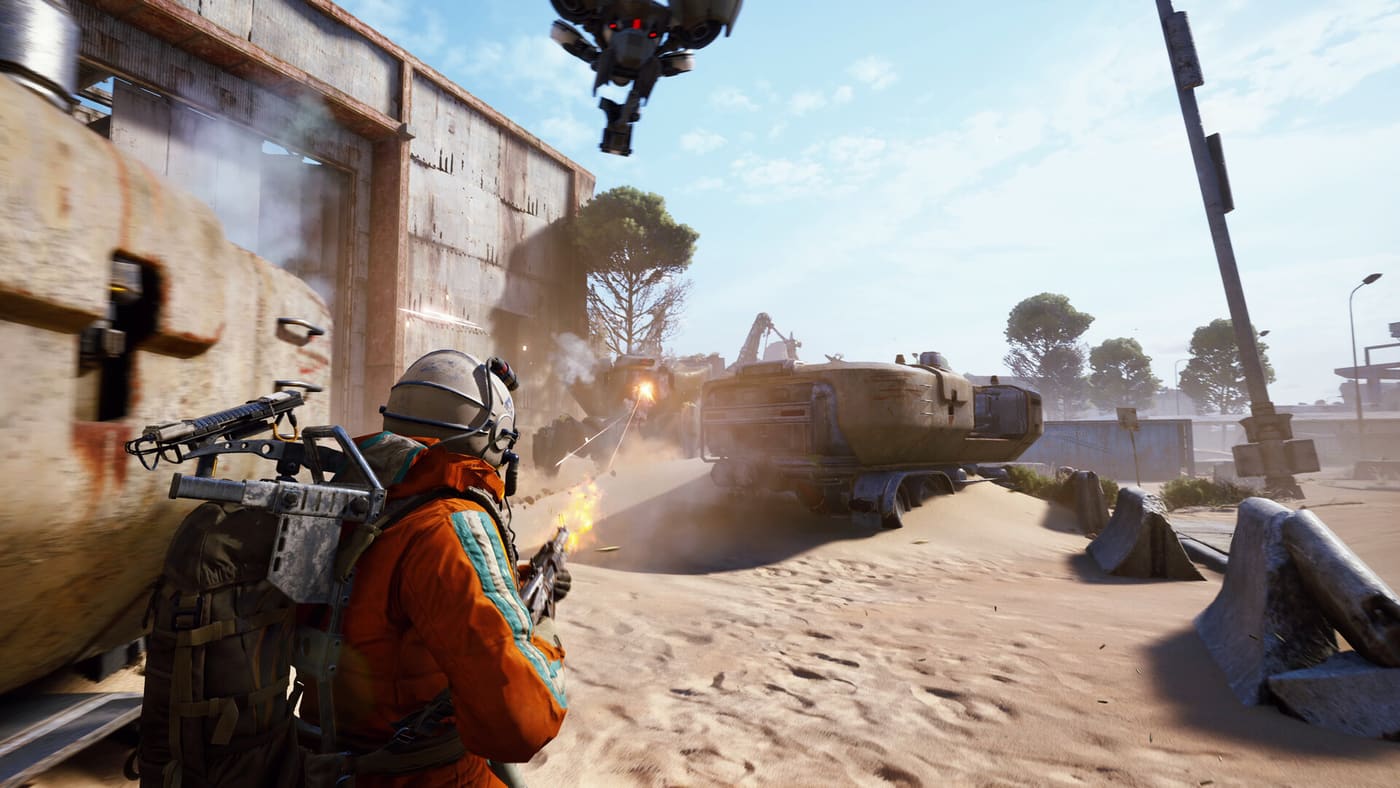 A Raider in an orange suit takes cover behind a rusted metal barrier as a huge tracked ARC machine grinds across open sand toward him and a smaller flying drone closes in overhead. Official ARC Raiders screenshot supplied by Embark Studios.