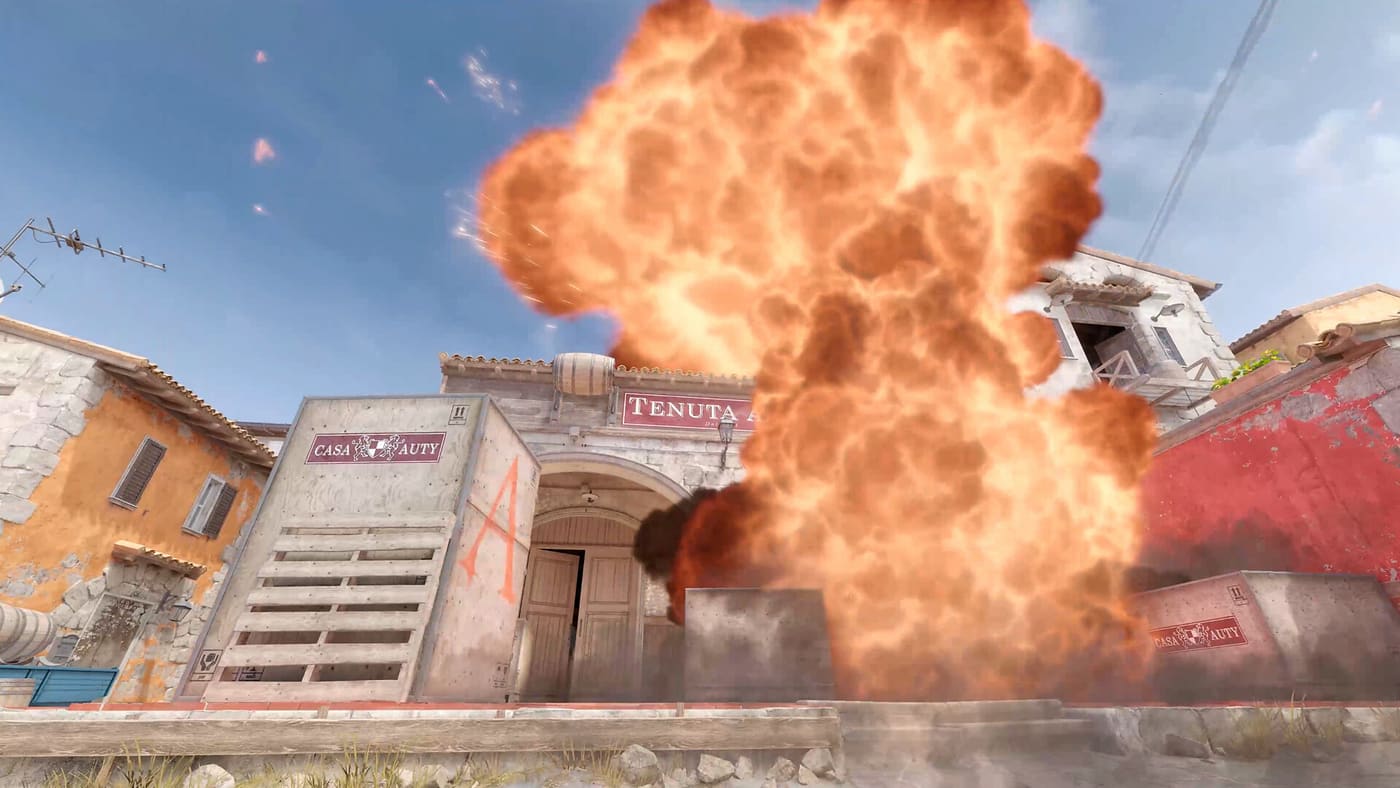 A bomb detonates at the A site on Inferno, a column of orange fire rising over the site building and the roofs behind it. Official Counter-Strike 2 screenshot supplied by Valve.
